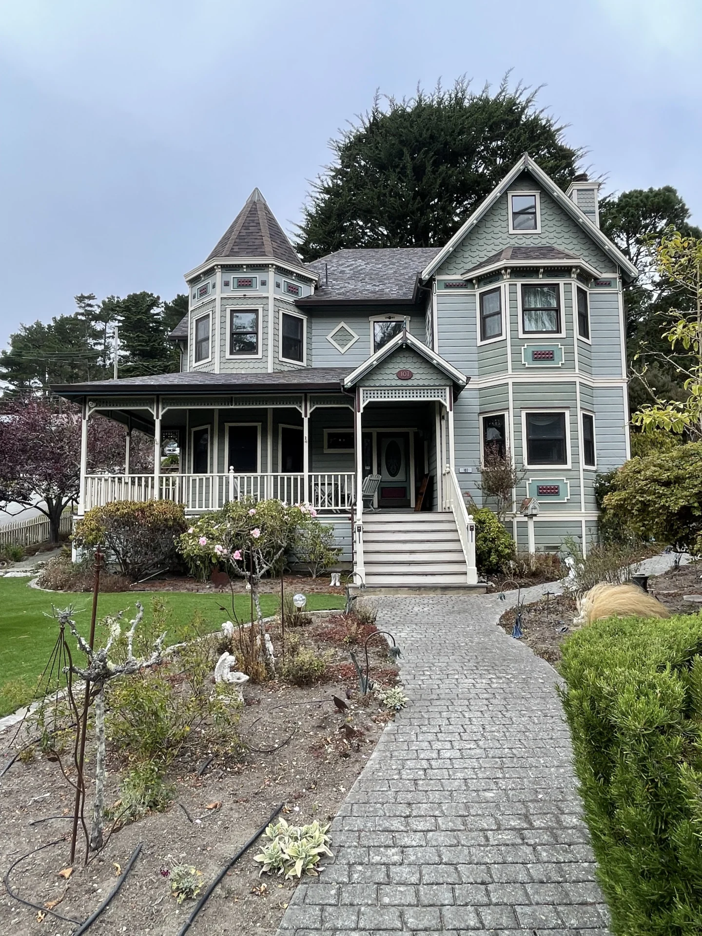 Victorian-style house with a turret, light green siding, and white trim. The house has a covered porch and a garden with a pathway leading to the front steps.